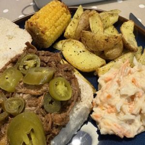 A barbecue pulled brisket roll sat on a plate with corn on the cob, wedges and coleslaw