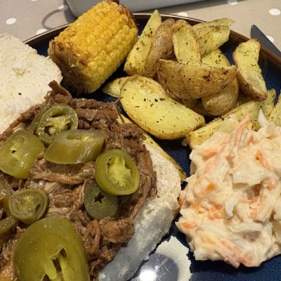 A barbecue pulled brisket roll sat on a plate with corn on the cob, wedges and coleslaw